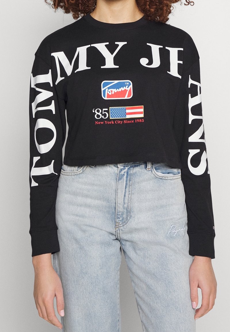 Black long-sleeve crop top with white text "TOMMY" and "MY JEANS" on sleeves. Features a logo and red, white, and blue design on the front.