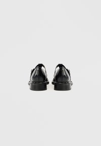 Μαύρα δερμάτινα παπούτσια με λεία επιφάνεια, στρογγυλεμένη μύτη και χαμηλό στρωμένο τακούνι· διαθέτουν λουράκι πίσω και λεπτομέρεια με πόρπη.