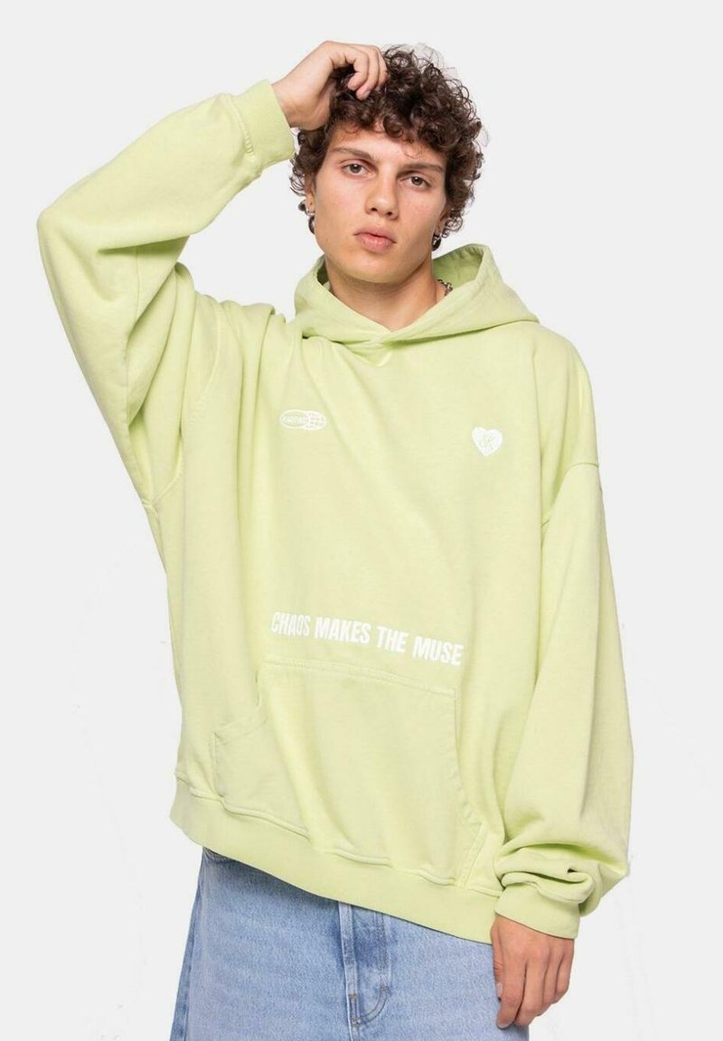Sudadera verde claro con un bolsillo frontal, puños acanalados y un cordón ajustable. Presenta un logo de corazón blanco y el texto "EL CAOS HACE A LA MUSA".