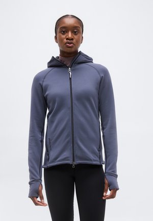 Frau trägt eine graue Kapuzenjacke mit Reißverschluss und Daumenlöchern, steht vor einem schlichten hellen Hintergrund.