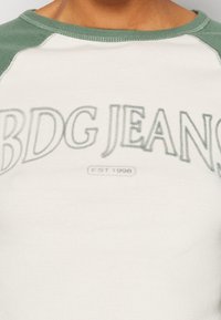 Světle šedé tričko s zelenými raglánovými rukávy. Obsahuje text "BDG JEANS" v mírně vyvýšené tmavší šedé barvě. Měkká bavlněná tkanina, volný střih.
