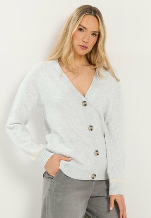 Blonde Frau trägt eine hellgraue, geknöpfte Strickjacke und graue Hose, steht mit einer Hand in der Tasche vor einem einfarbigen Hintergrund.