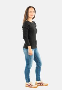 Donna in piedi di lato che indossa un maglione nero, jeans blu e sneakers beige con strisce bordeaux su uno sfondo bianco.