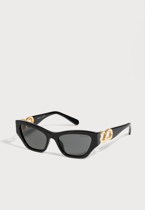 Lunettes de soleil noires œil-de-chat, avec des verres gris foncé et des détails dorés sertis de cristaux sur les branches, sur un fond clair.