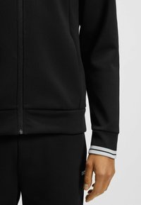 Felpa nera con zip, caratterizzata da una texture liscia, colletto alto e polsini a righe. Il design include una zip frontale e una vestibilità slim.