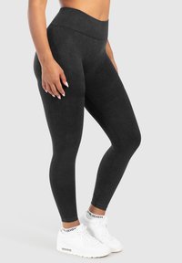 Vrouw in hoge zwarte leggings en witte sportschoenen, staand tegen een effen lichte achtergrond.