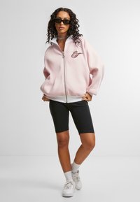 Felpa rosa chiaro con zip e colletto bianco, con un logo sul petto. Completa il look un paio di short da bicicletta neri e sneakers bianche.