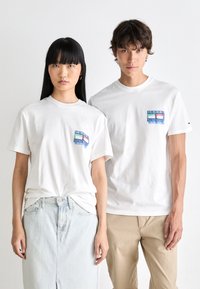 Camisetas de algodón blancas con un parche de logo colorido en el lado izquierdo del pecho, corte relajado, con mangas cortas y diseño de cuello redondo.