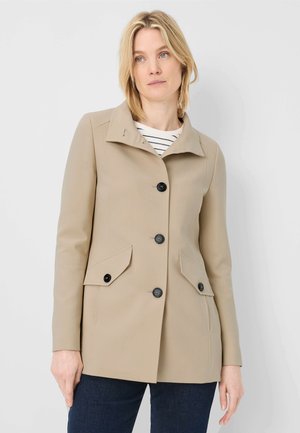 Frau trägt einen beigen, geknöpften Mantel mit Pattentaschen und eine marineblaue Hose, steht vor einem einfarbigen, hellen Hintergrund.