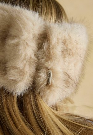 Bēšas pūkainas ausu aizsargu austiņas pār gariem blondiem matiem ar redzamu nelielu taisnstūrveida "Joules" zīmola etiķeti.