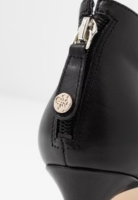 Bottine en cuir noir avec un bout pointu, un talon haut, une fermeture éclair latérale avec quincaillerie argentée, et un détail décoratif de bouton embossé.