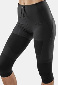 Schwarze Kompressions-Capris aus glattem Material, ergonomischer Form, mit strukturierten Akzenten und einem Kordelzugbund. CEP-Logo sichtbar.