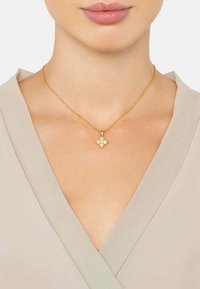 Kultaketju, jossa on kukkaketju ja karhea pinta sekä kolme valkoista kiveä, käytetty paljaalla kaulalla beigessä paidassa.