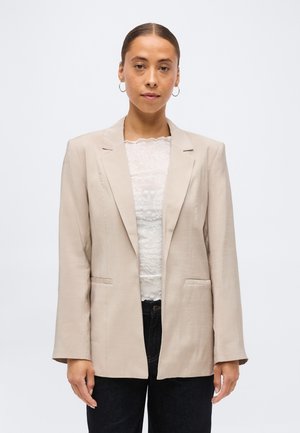 Femme aux cheveux attachés, portant un blazer beige sur un haut en dentelle blanche et un pantalon noir, debout devant un fond clair uni.