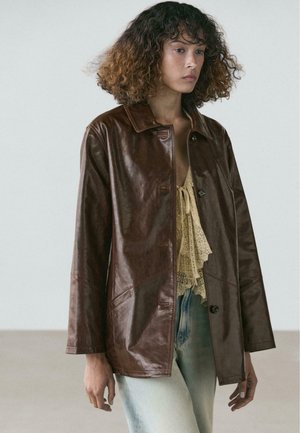 Jeune femme aux cheveux bouclés porte une veste en cuir marron foncé par-dessus un haut en dentelle crème et un jean bleu clair, debout devant un fond uni.