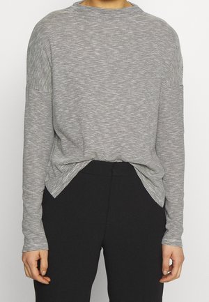 Personne portant une chemise à manches longues, fine, rayée noir et blanc, légèrement rentrée dans un pantalon noir taille haute, les mains détendues le long du corps.