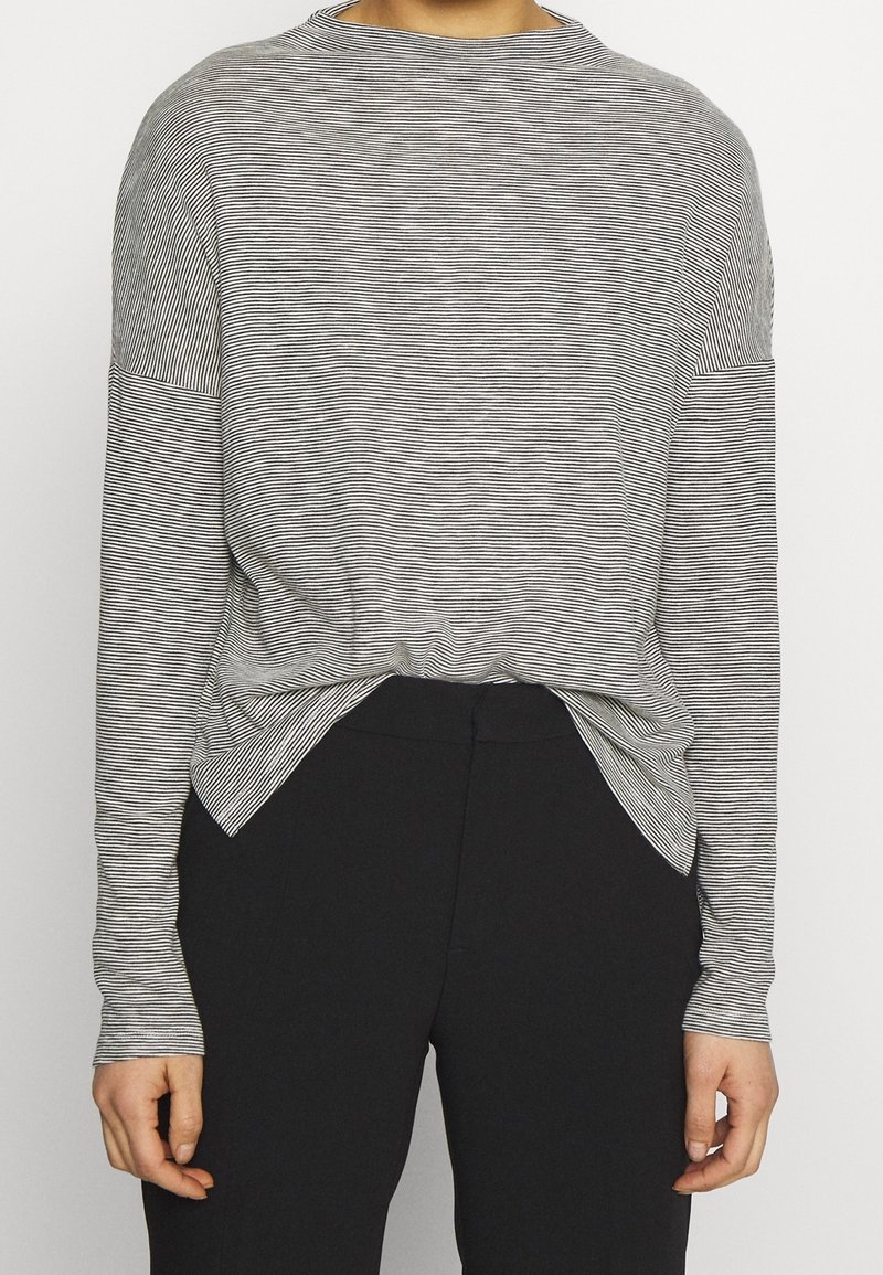 Personne portant une chemise à manches longues, fine, rayée noir et blanc, légèrement rentrée dans un pantalon noir taille haute, les mains détendues le long du corps.