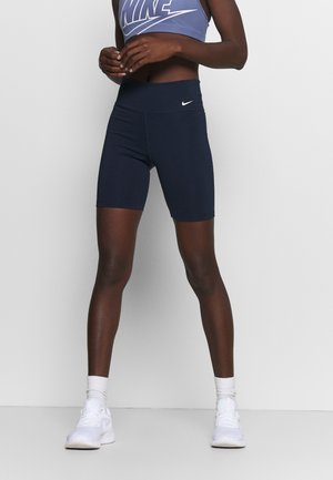 Fit persoon die donkerblauwe Nike-shorts, een blauwe Nike sportbeha, witte sokken en witte sportschoenen draagt, staand tegen een eenvoudige grijze achtergrond.