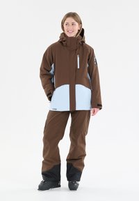 Braune und hellblaue Skijacke mit Kapuze, zwei Reißverschlusstaschen, kombiniert mit braunen Hosen und schwarzen Stiefeln. Hergestellt aus einem wasserdichten Material.