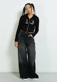 Czarna krótka bluza z zamkiem, na której widnieje liczba 13 w postarzanym nadruku, zestawiona z szerokimi, wyblakłymi czarnymi dżinsami i czarnymi trampkami.
