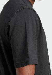 Man in een eenvoudig zwart T-shirt met korte mouwen, met gedetailleerde stiksels op de schouder en mouwnaad.