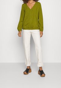 Blusa de manga larga verde oliva con escote en V y dobladillo elástico, decorada con pequeños puntos. Combinada con pantalones blancos remangados y zuecos negros.