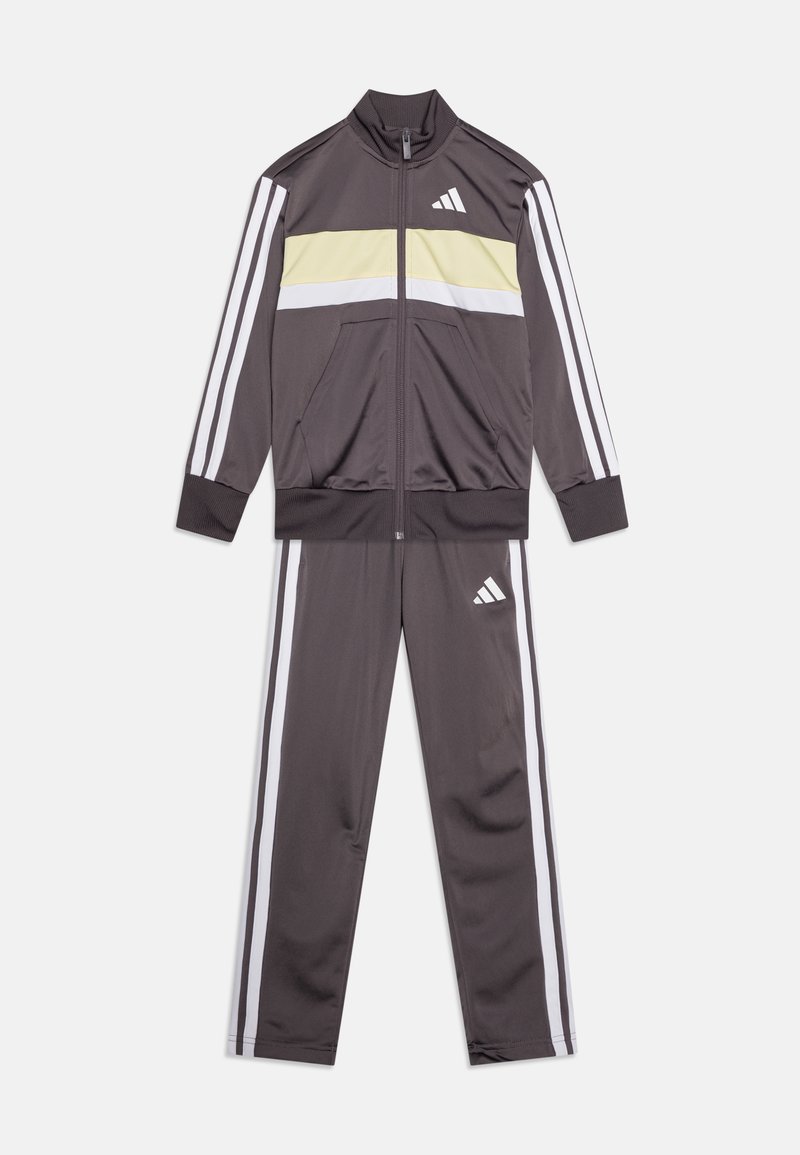 Γκρι φόρμα που χαρακτηρίζεται από ένα ζακέτα με φερμουάρ με κίτρινη και λευκή οριζόντια ρίγα, σε συνδυασμό με παντελόνι που ταιριάζει, και τα δύο με λευκές ρίγες στα πλάγια.