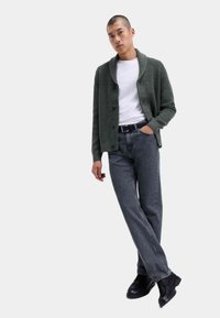 Grøn strikket cardigan, hvid t-shirt, grå jeans, sort bælte og sorte støvler. Cardiganen har en sjalskrave og knaplukning.