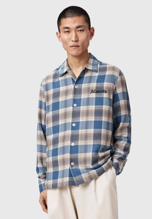 Mand iført blå og beige ternet skorte med knapper og "Allsaints"-logo, kombineret med lyse bukser, stående mod en ensfarvet baggrund.