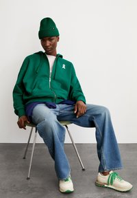 Grüne Zip-Hoodie mit weißem Bändchen-Logo, kombiniert mit geschichteten blauen und weißen Oberteilen, weiten Jeans und Sneakern mit grünen Akzenten.