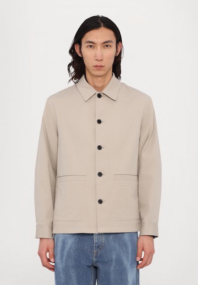 Veste en coton beige avec des boutons noirs, un col pointu et deux poches avant. Portée sur un jean bleu clair, elle présente une coupe droite et un design minimal.
