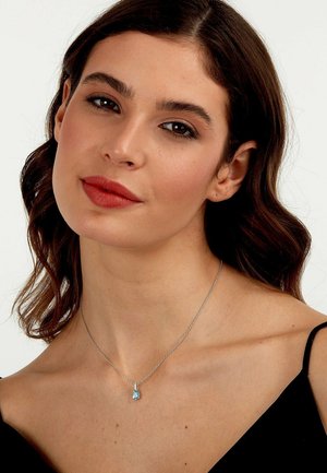 Silberkette mit einem tropfenförmigen blauen Edelsteinanhänger, der auf einem nackten Schlüsselbein ruht, vor einem schwarzen Hintergrund.