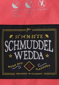 Röd vattentålig tyg med en svart etikett med vit och guld text som säger "SCHMUDDEL WEDDA" samt symboler för vattentåliga och vindskyddande egenskaper.