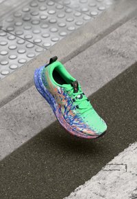 Zapatilla deportiva verde brillante con patrones multicolores que pasan a acentos en azul y naranja, con una suela texturizada y detalles de marca en negro.