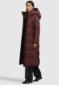 Langer, burgunderfarbener Puffercoat mit großer Kapuze. Verfügt über seitliche Reißverschlüsse, schwarze Details, diagonale Nähte und ein Logo am Ärmel.