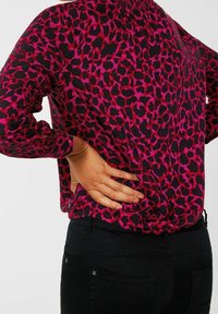 Rosa-schwarz gemustertes Leopardenoberteil mit langen Ärmeln, gerafftem Halsausschnitt und lockerem Sitz; kombiniert mit schwarzen Hosen.