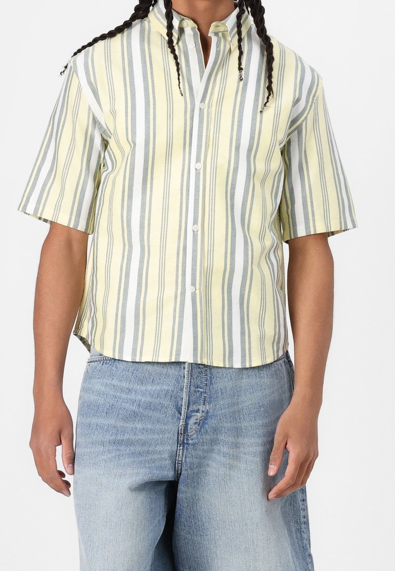 Personne portant une chemise à manches courtes boutonnée avec des rayures verticales jaunes, blanches et grises et un jean en denim bleu clair.