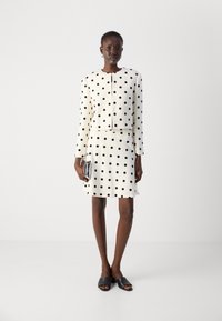 White polka dot jacket and skirt set featuring long sleeves, round neckline, textured fabric, and a straight cut. Black slide sandals and clutch.
