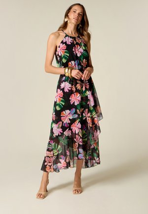 Frau in einem ärmellosen schwarzen, floralen Neckholderkleid mit bunten pinken und grünen Blättern, trägt beigefarbene Absätze und goldene Armbänder, steht vor schlichtem Hintergrund.