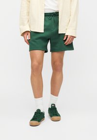 Πράσινα βαμβακερά σορτς με κορδόνι, σετ με λευκές κάλτσες με μανσέτες και πράσινα αθλητικά παπούτσια με καφέ σόλα. Επάνω, ένα ανοιχτόχρωμο πουκάμισο με κουμπιά.