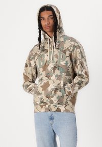 Kamouflage hoodie i beige, brunt och teal med framficka och dragskohuva; har djärv "GAP" tryck över bröstet.