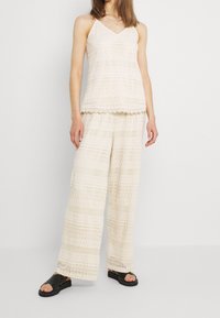 Completo in maglia di pizzo composto da top e pantaloni a gamba larga in un delicato beige, caratterizzato da motivi testurizzati e bordi scallop, abbinato a sandali neri.