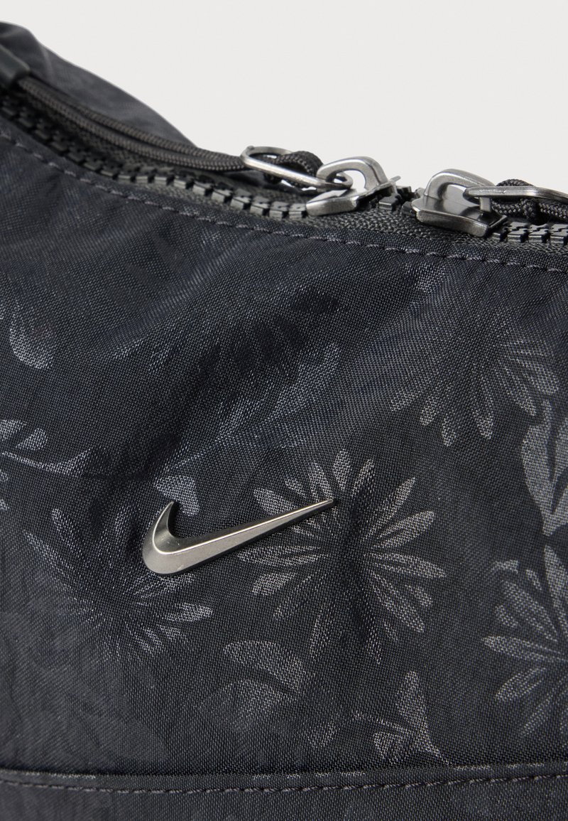 Nahaufnahme von schwarzem Stoff mit dezentem Blumenmuster, metallischem Nike-Logo und zwei Reißverschlüssen an einer Tasche oder einem Beutel.