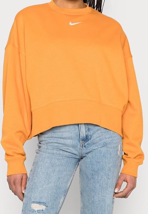 Personne portant un sweat-shirt Nike orange ample avec un logo Swoosh blanc et un jean bleu clair déchiré, debout devant un fond uni.