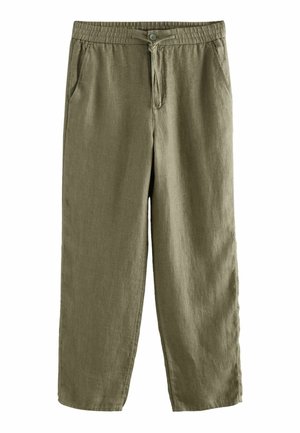 Die rechte Hand ruht neben olivgrüner Leinenhose mit sichtbarer Gesäßtasche und Bund unter einem weißen Hemd.