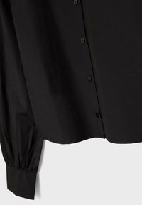 Camicia nera a maniche lunghe con polsini arricciati e bottoni visibili lungo la patta frontale, mostrata su uno sfondo semplice.