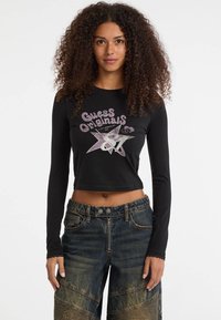 Schwarzes, langärmliges, kurzes Oberteil mit "Guess Originals" Grafik in Pink und Silber, kombiniert mit blauen, ausgewaschenen Denim-Hosen mit strukturierten Akzenten.