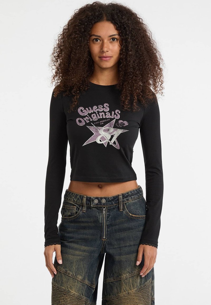 Zwarte cropped longsleeve shirt met "Guess Originals" grafiek in roze en zilver, gecombineerd met blauw vervaagde denim broek met textuurelementen.
