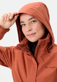 Giacca impermeabile arancione con cappuccio aderente, zip visibili e logo sul petto. Texture liscia e design attivo per l'uso all'aperto.