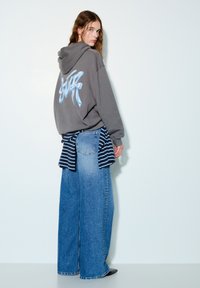 Siva dukserica s plavom grafikom na leđima, u kombinaciji s velikim plavim trapericama. Navy majica na pruge vezana oko struka.